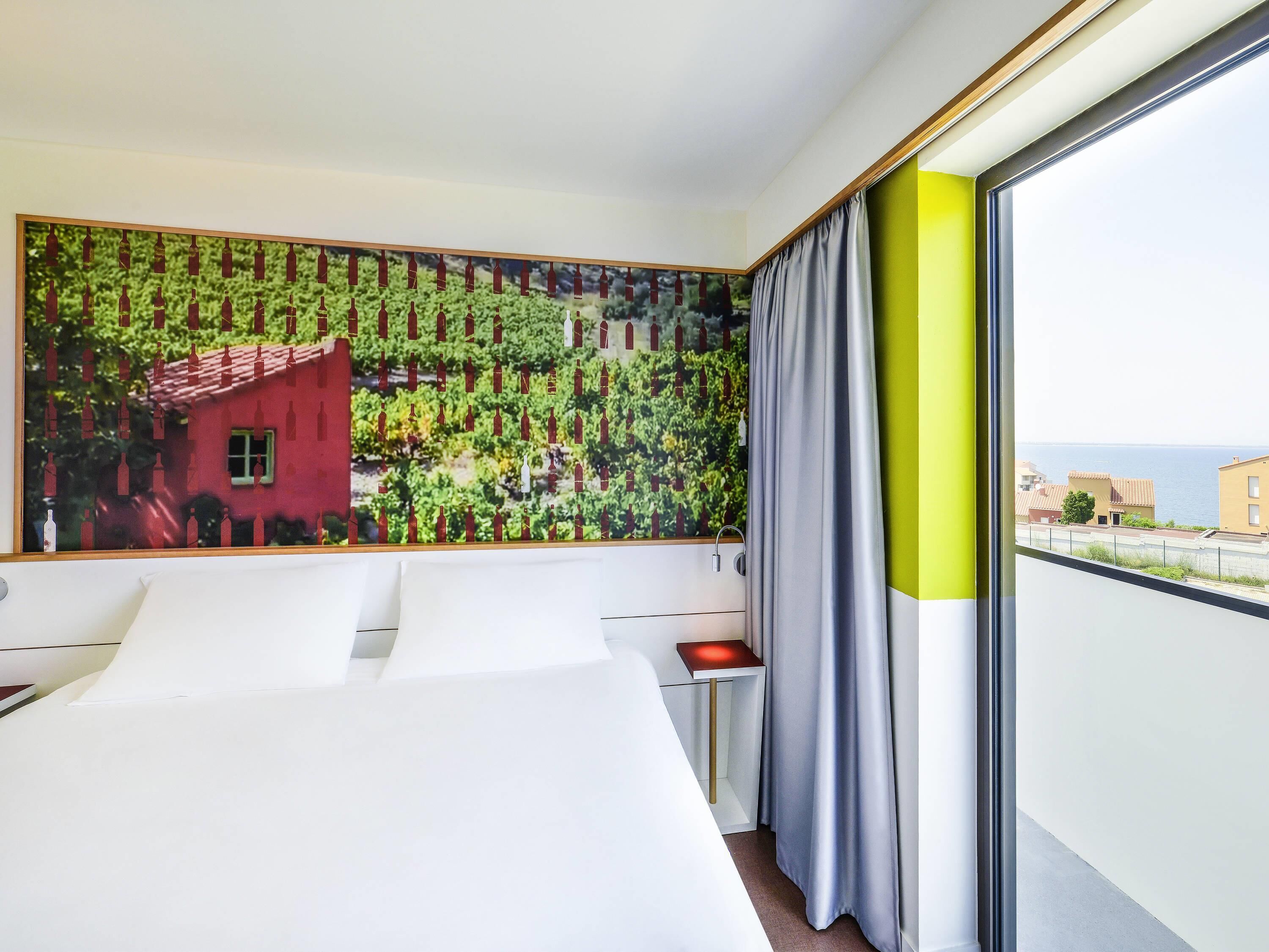 A hotel room with a large window, a bed, a bedside table, and a wall-mounted artwork.