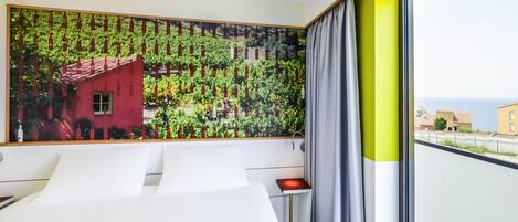 A hotel room with a large window, a bed, a bedside table, and a wall-mounted artwork.