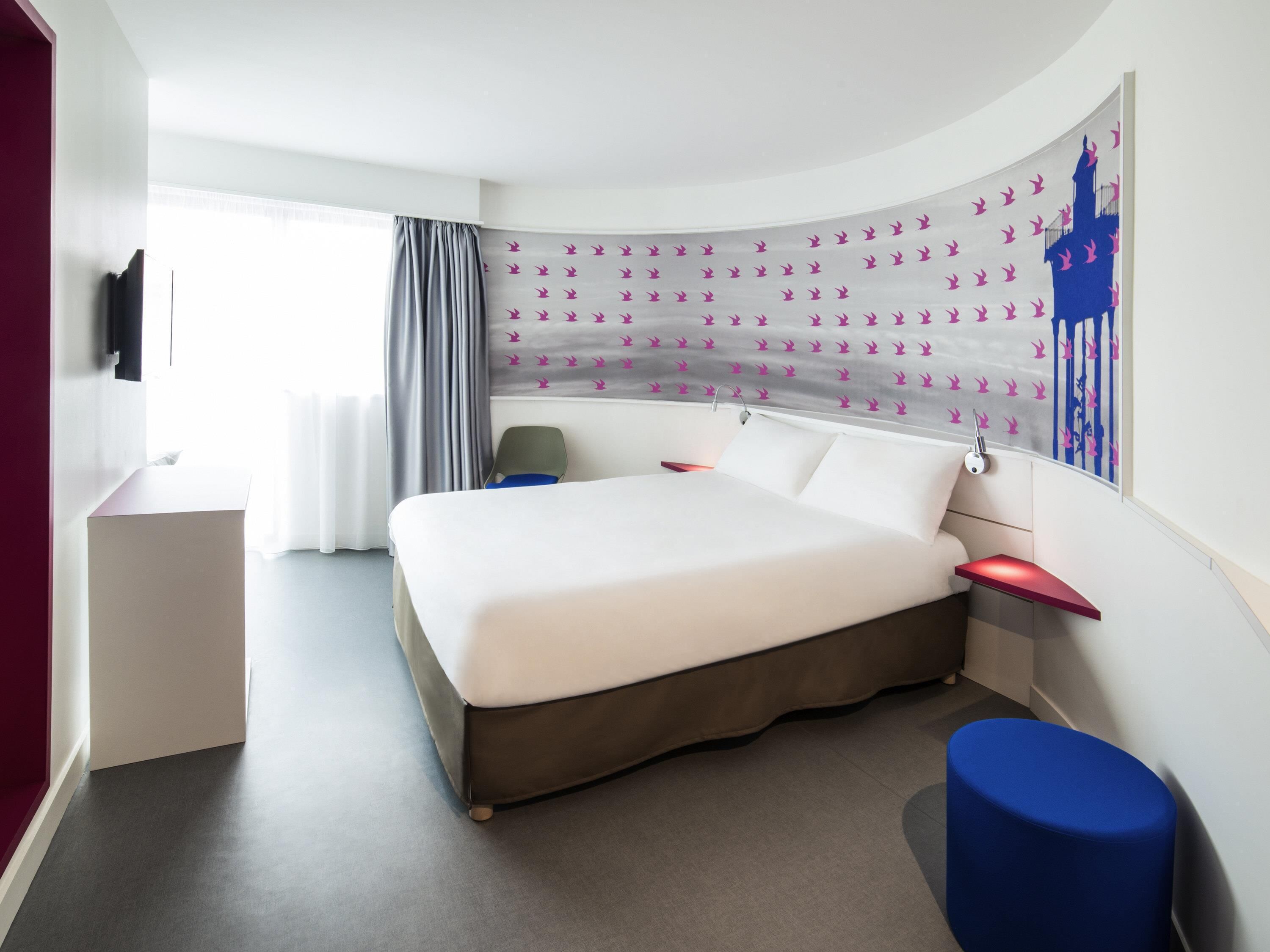 Une chambre d’hôtel moderne avec un lit aux lignes courbes, un bureau, une chaise et une grande fenêtre avec des rideaux.