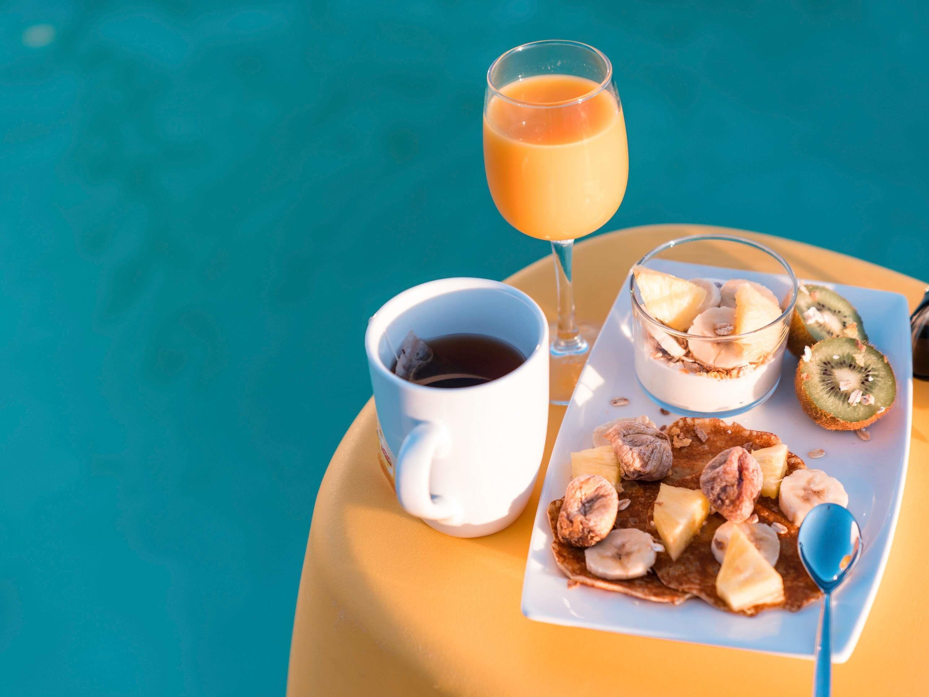 Un petit-déjeuner composé d’une tasse de café, d’un verre de jus d’orange, d’un bol de yaourt aux fruits et d’une assiette de crêpes et de bananes.