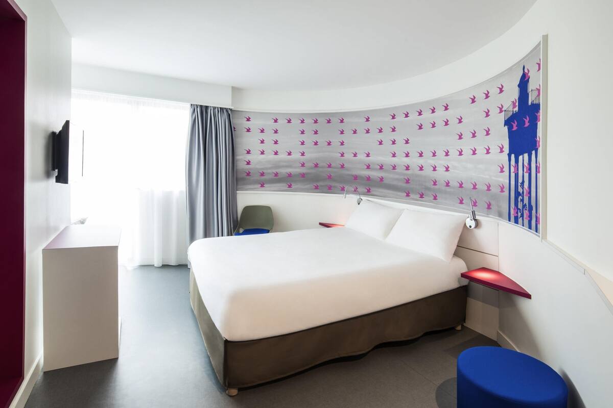 Une chambre d’hôtel moderne avec un lit aux lignes courbes, un bureau, une chaise et une grande fenêtre avec des rideaux.