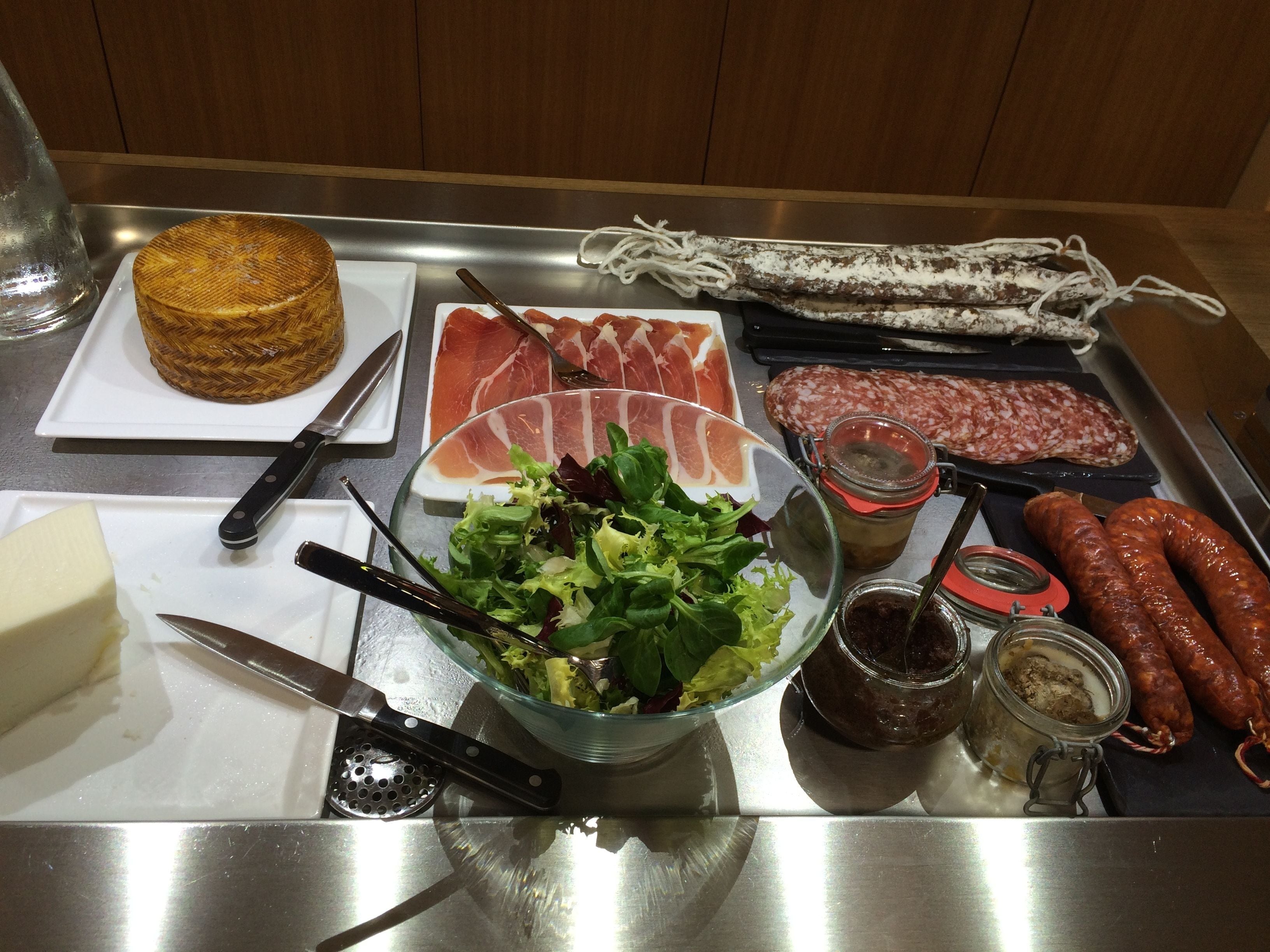 Una mesa de acero inoxidable con diversos alimentos, incluyendo fiambres, ensalada, queso y salsas.