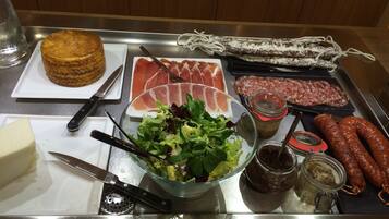 Una mesa de acero inoxidable con diversos alimentos, incluyendo fiambres, ensalada, queso y salsas.