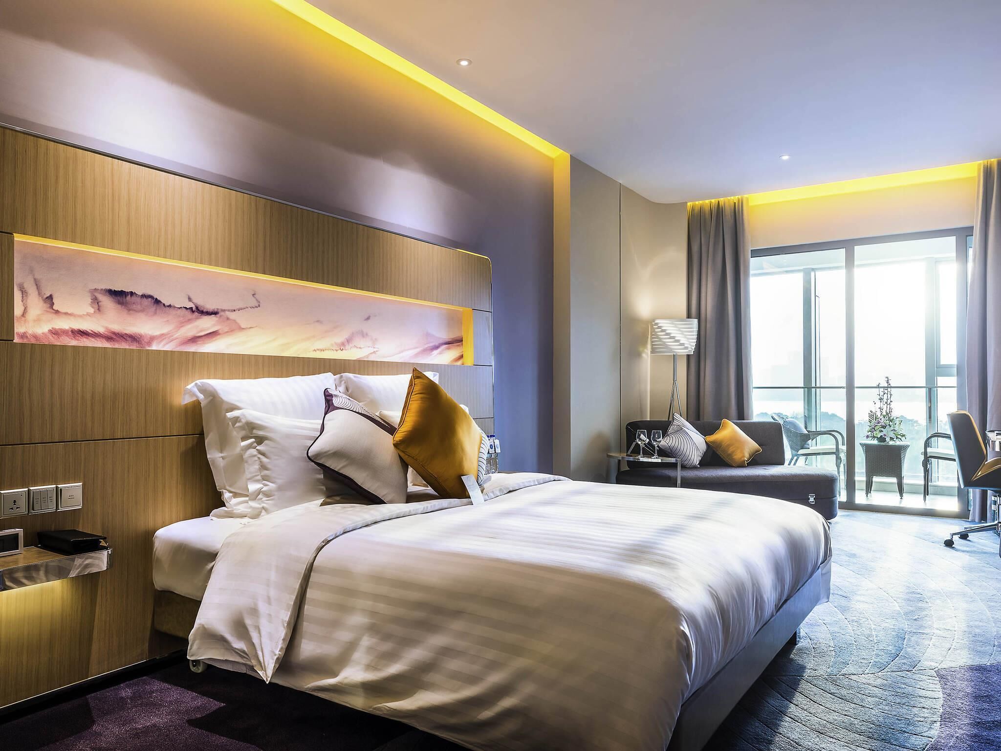 A modern hotel room with a large bed, a desk, a chair, and a view of the outdoors.