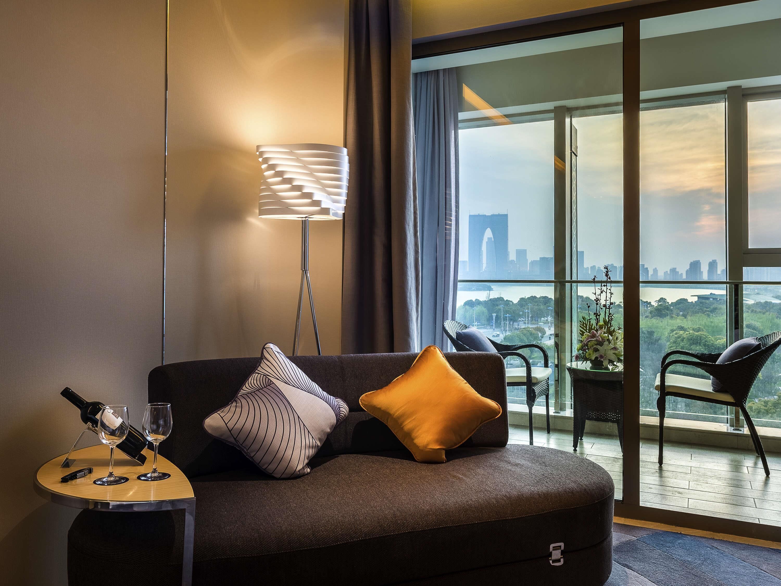 A modern hotel room with a sofa, a floor lamp, a small table with wine glasses, and a view of a cityscape through large windows.