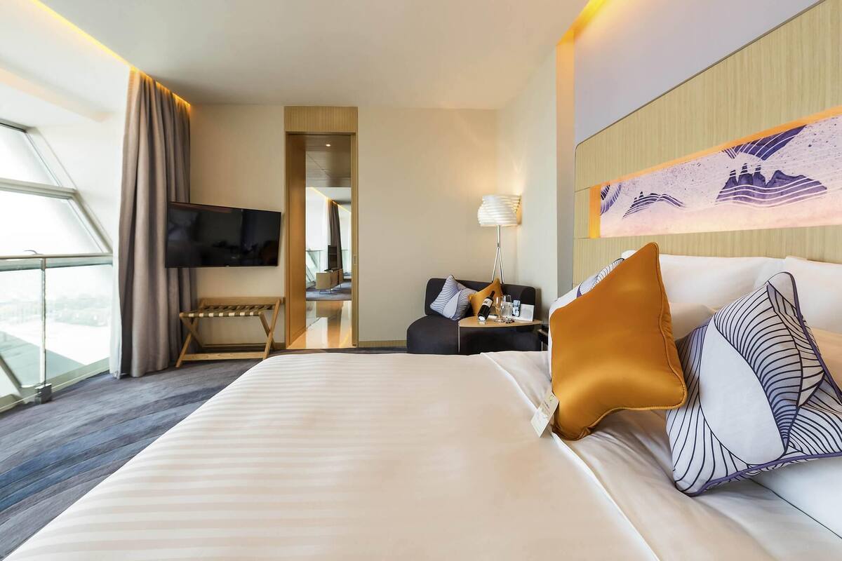 A hotel room with a large bed, a TV, a desk with a lamp, and a view through a window.
