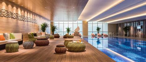 A modern indoor pool area with wooden flooring, seating arrangements, and a large window providing natural light.