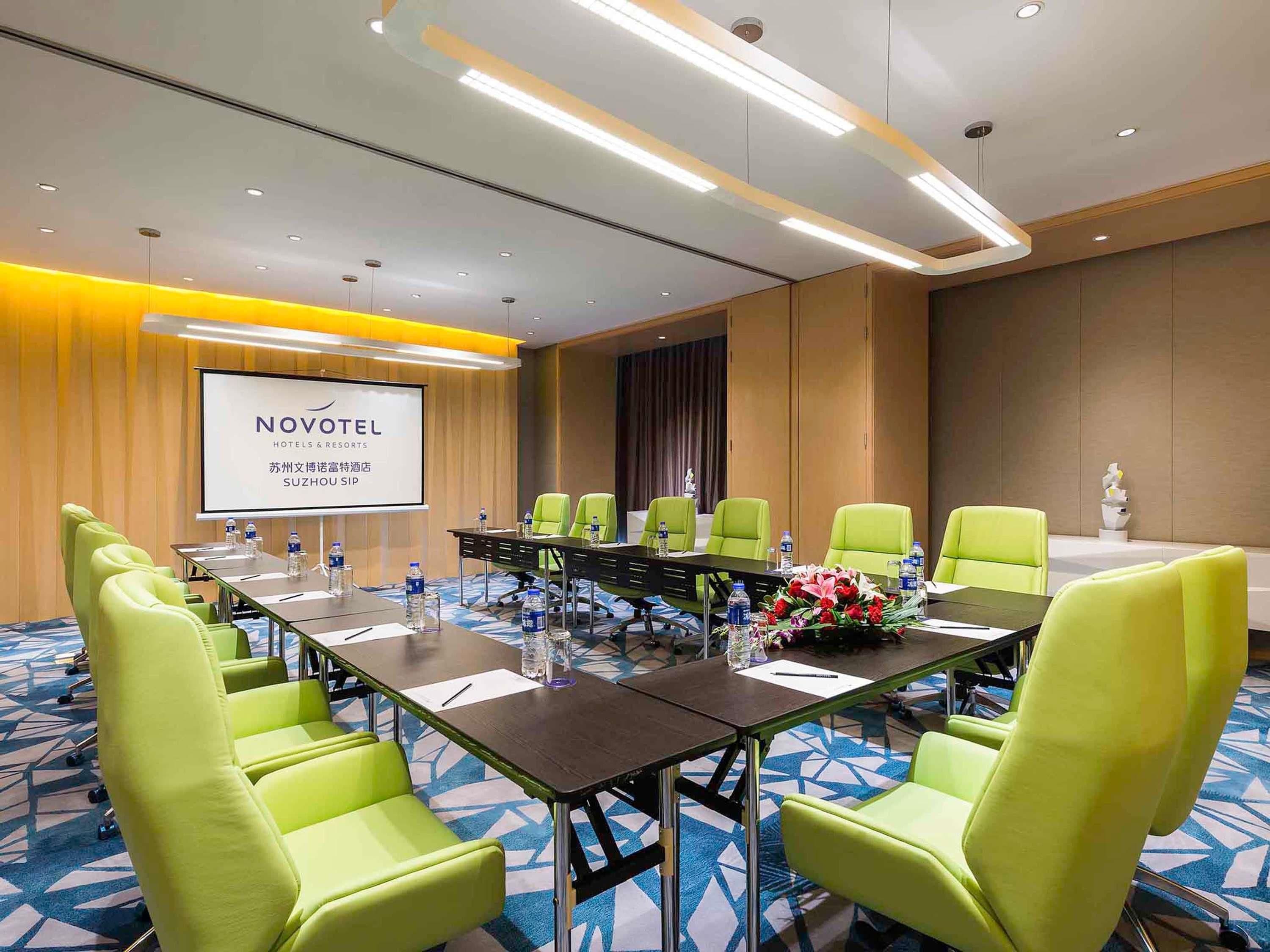 Un salón de conferencias con una larga mesa, sillas verdes y una pantalla de proyector que muestra la marca NOVOTEL.