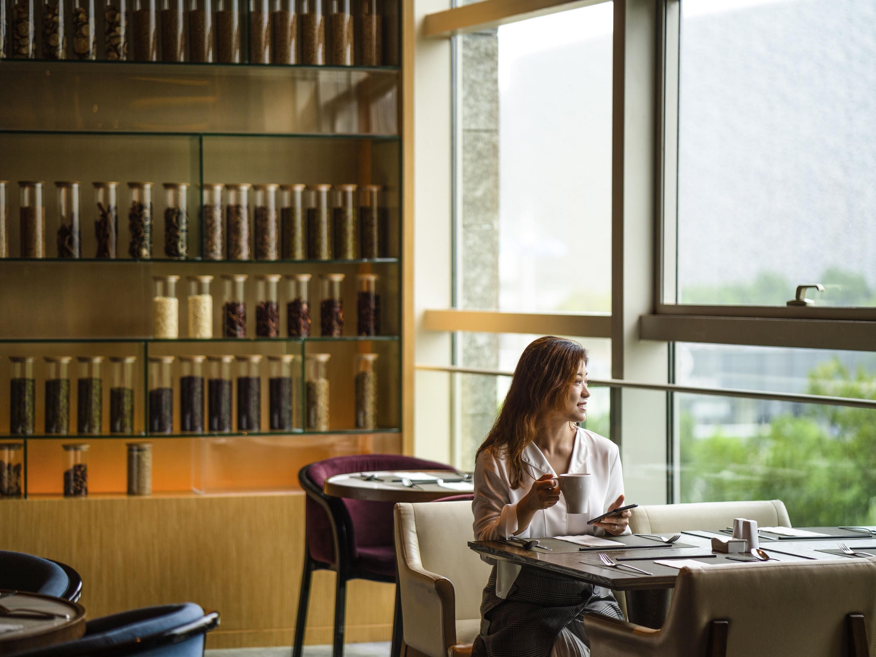 A person seated at a table with a smartphone and a cup, in a room with a large window and a bookshelf filled with jars.