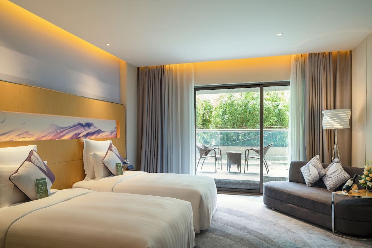 A hotel room with two beds, a sofa, a balcony with outdoor seating, and a painting on the wall.