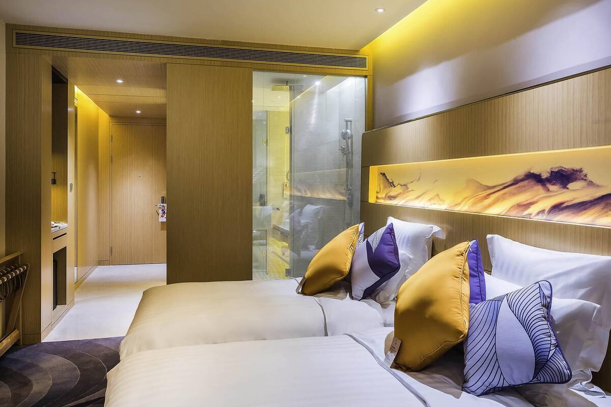 A modern hotel room with two beds, a large window with blinds, a wall-mounted artwork, and a wooden door.