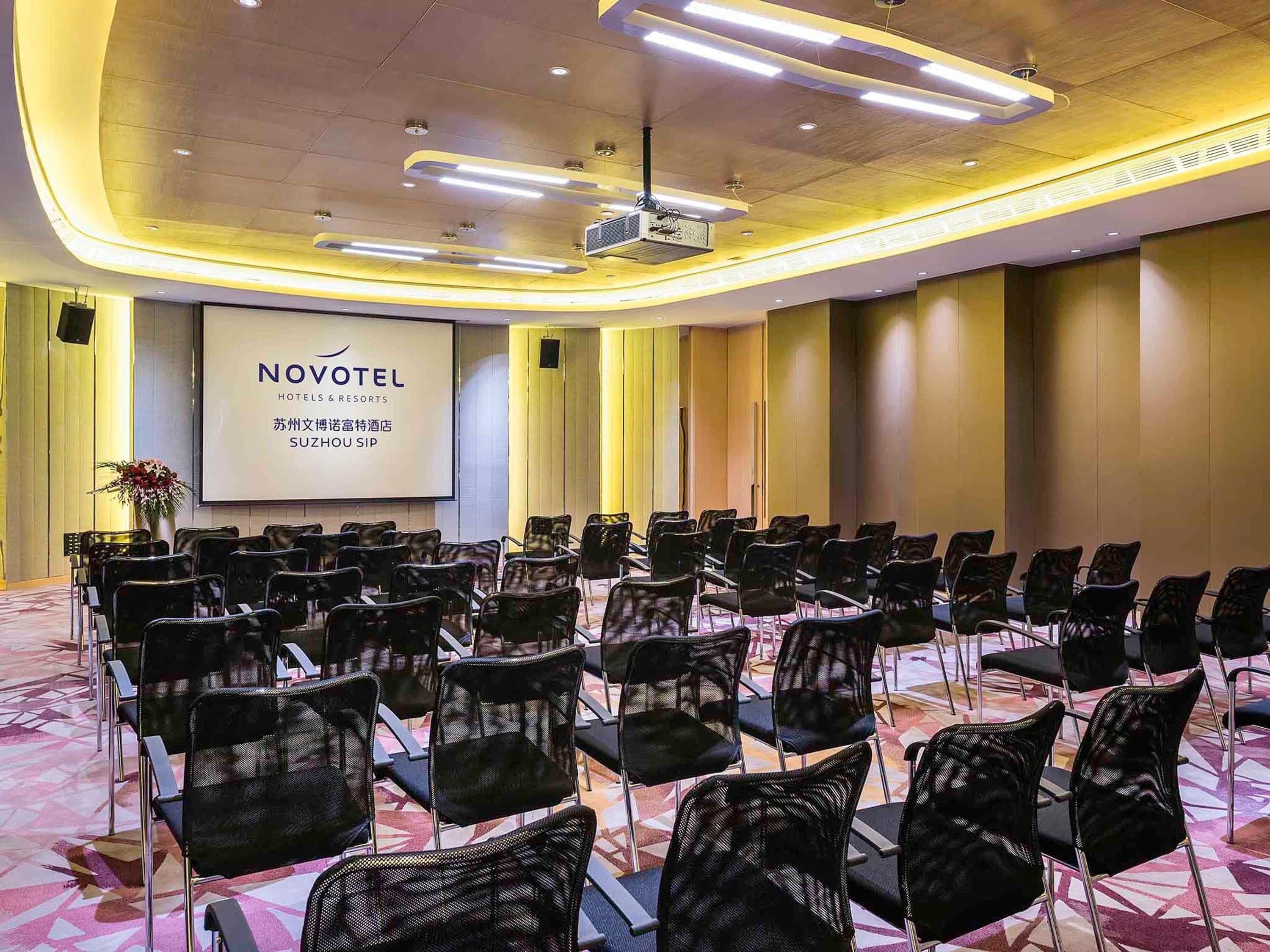 A conference room with rows of chairs, a projector screen, and a large screen displaying 'NOVOTEL HOTEL & RESORTS' text.