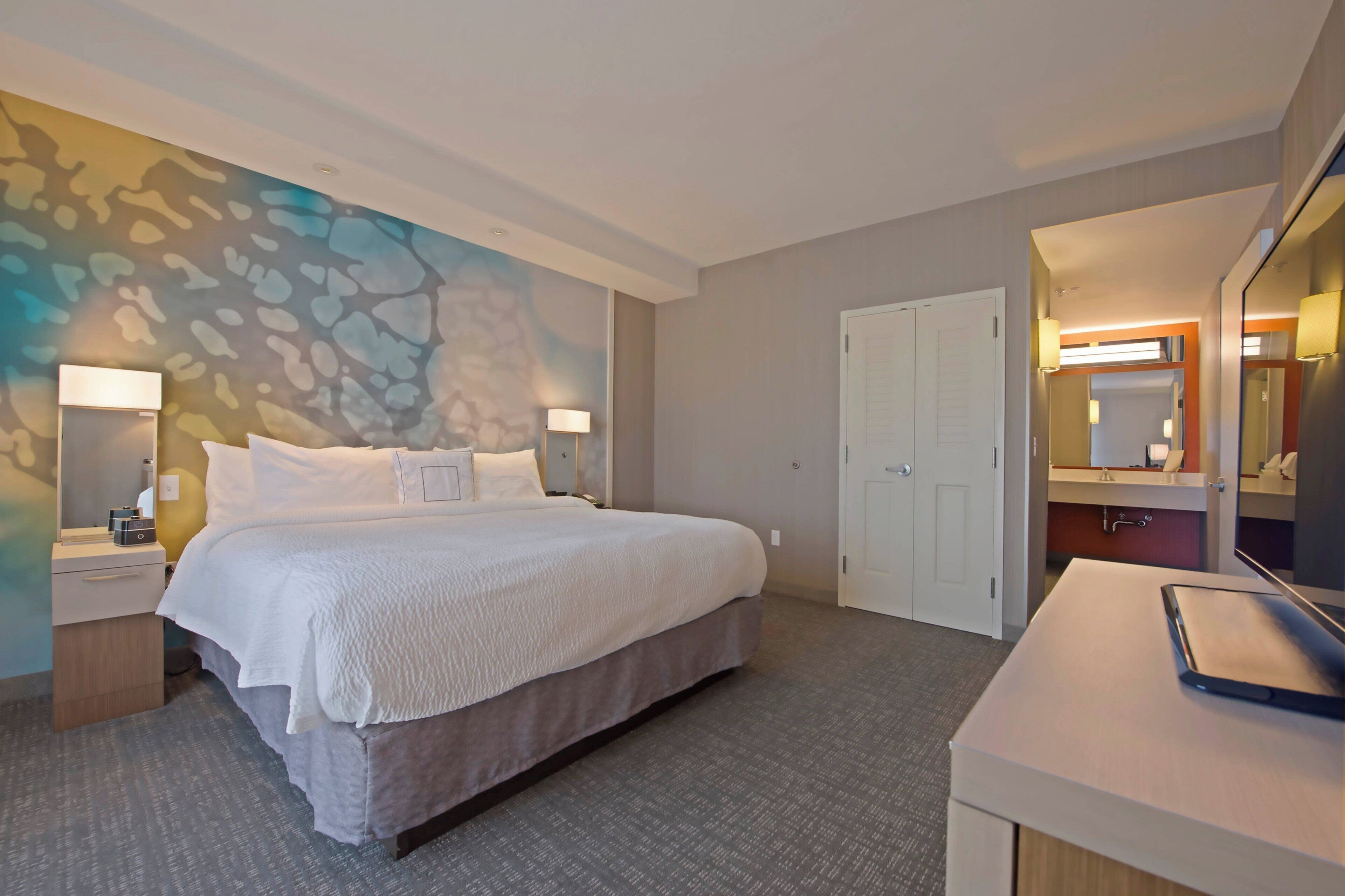 A hotel room with a large bed, a desk with a laptop, a mirror, and a door leading to a bathroom.