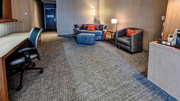 A hotel room with a desk, office chair, sofa, and armchair. There is a lamp, a coffee machine, and a small table with a remote control.