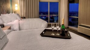Een hotelkamer met twee bedden, een nachtkastje, een dienblad met champagneglazen en een fles, en een uitzicht op de stad in de schemering.