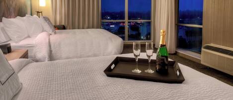 Habitación de hotel con dos camas, una mesita de noche, una bandeja con copas de champán y una botella, y vistas a la ciudad al anochecer.