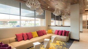 A modern lounge area with a sectional sofa, multiple cushions, and geometric side tables. Large windows offer views of the outside.