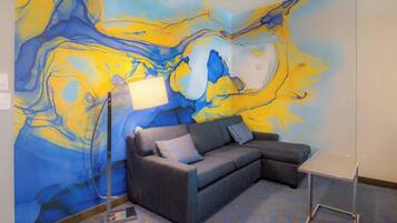 Une pièce décorée d’une fresque murale abstraite aux couleurs bleu et jaune, avec un canapé gris orné de coussins à rayures et une petite table blanche.