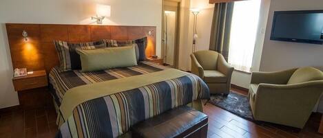Una habitación de hotel con una cama grande, dos sillones, un televisor de pantalla plana y una mesita de noche con lámpara.