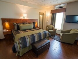Una habitación de hotel con una cama grande, dos sillones, un televisor de pantalla plana y una mesita de noche con lámpara.
