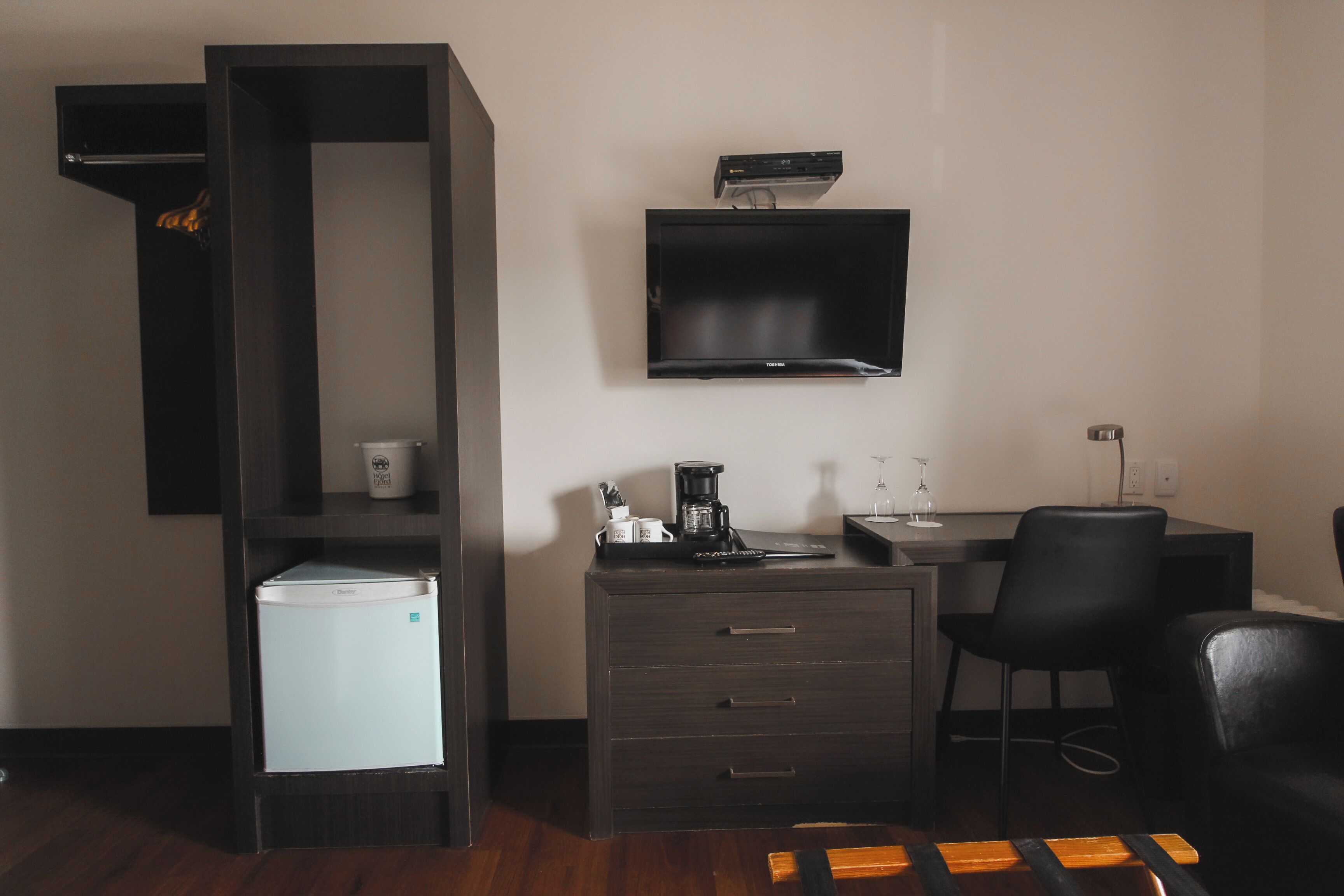 Une chambre d’hôtel compacte dotée d’une kitchenette, d’un téléviseur à écran plat fixé au mur, d’un petit réfrigérateur et d’un bureau avec une chaise.