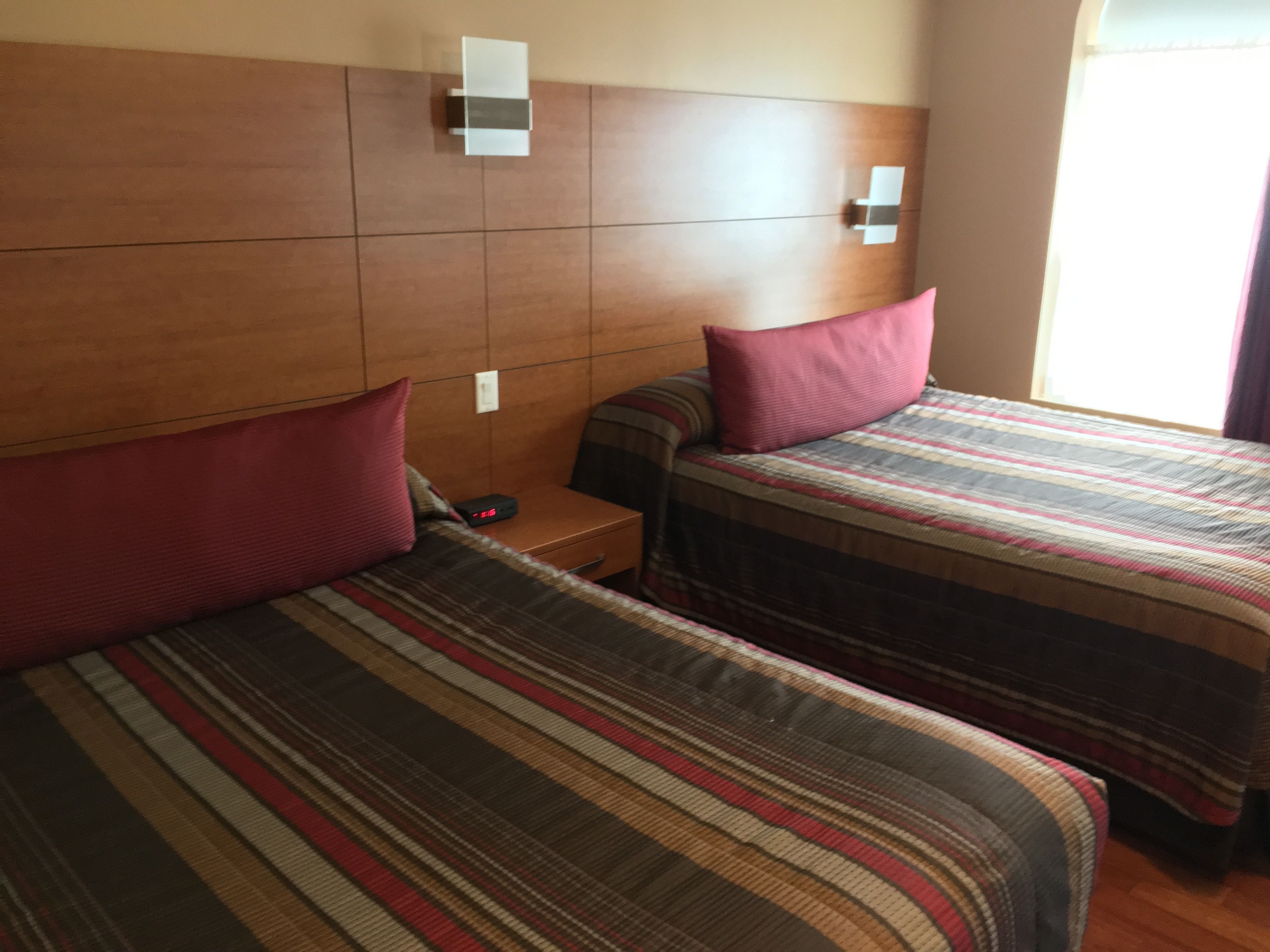 Une chambre d’hôtel avec deux lits, chacun doté de draps à rayures et d’un oreiller. Une table de chevet avec un téléphone et une lampe se trouve entre les deux lits. La chambre est équipée d’une tête de lit en bois et d’une fenêtre avec des rideaux.