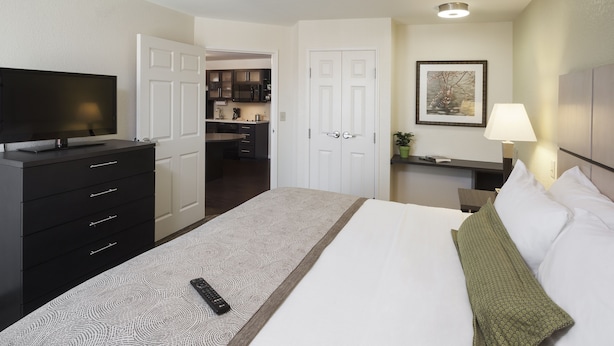 Habitación de hotel con cama, cómoda, televisión, escritorio con lámpara y vista hacia la cocina.