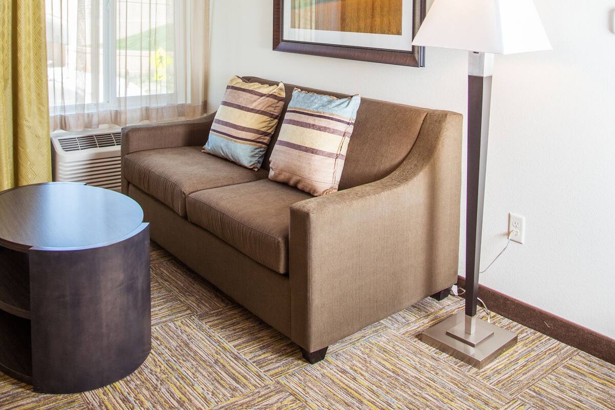 A hotel room with a brown sofa, a round table, a lamp, and a window with curtains.