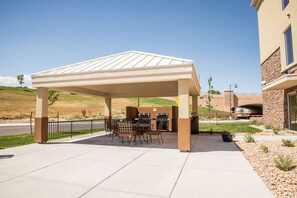 An outdoor patio with a covered seating area, barbecue grills, and a stone building in the background. - Candlewood Suites Denver North - Thornton by IHG (Thornton)