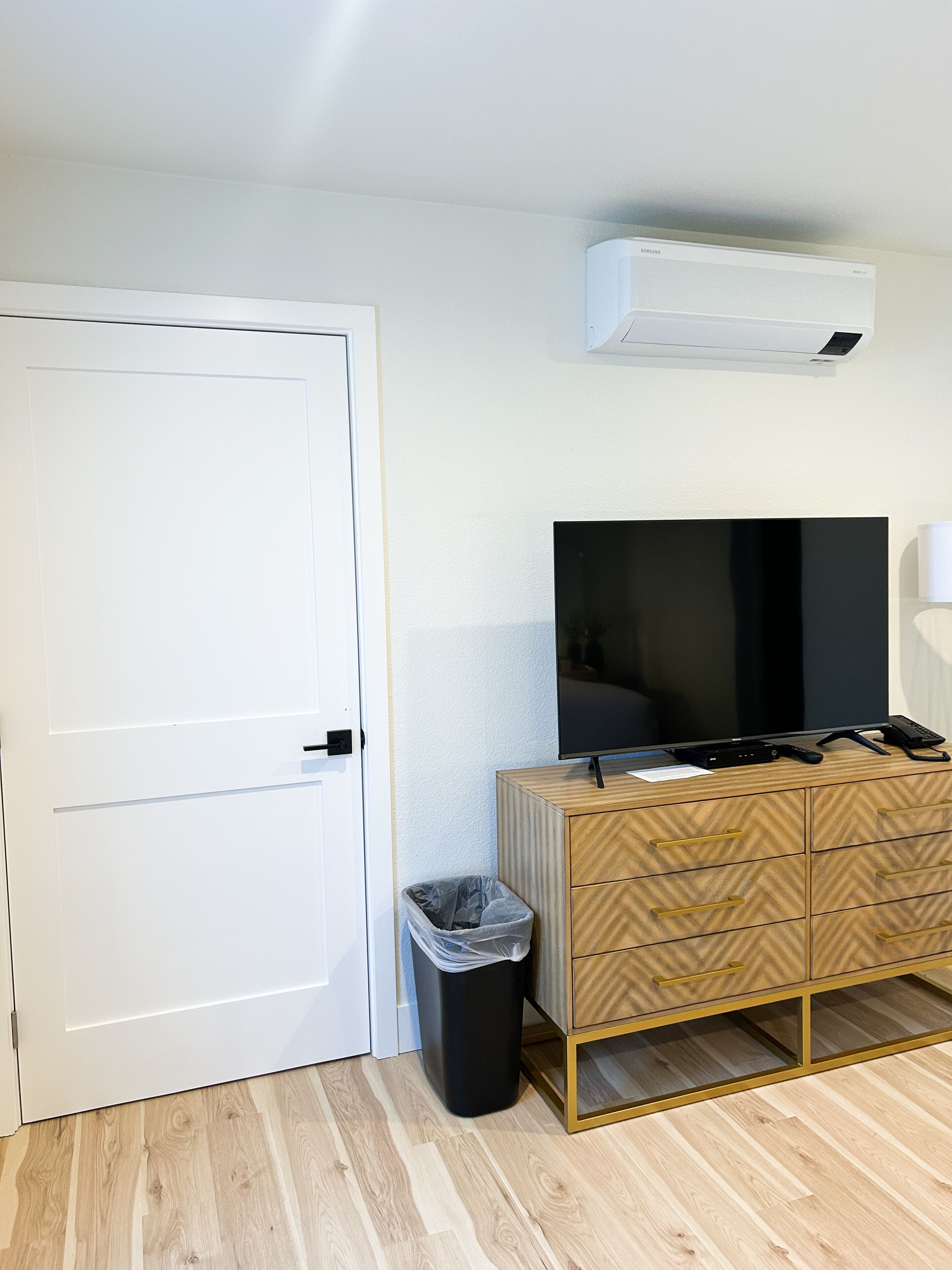 Ein Zimmer mit weißer Tür, einem Holzkasten mit Schubladen, einem schwarzen Mülleimer und einer fest installierten Klimaanlage.
