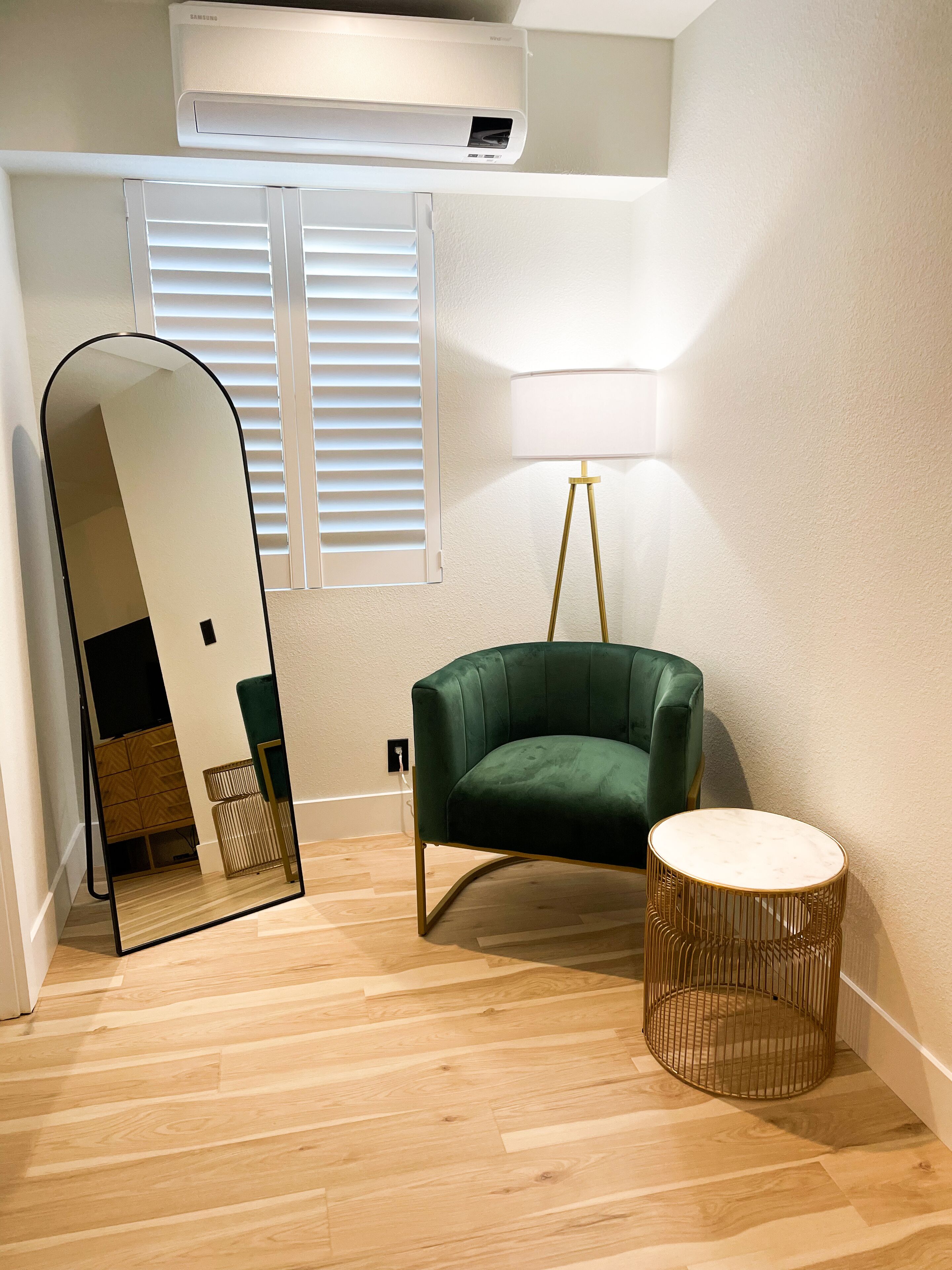 Una habitación con un sillón verde, una mesita redonda, una lámpara de pie, un espejo grande y una ventana con persianas.