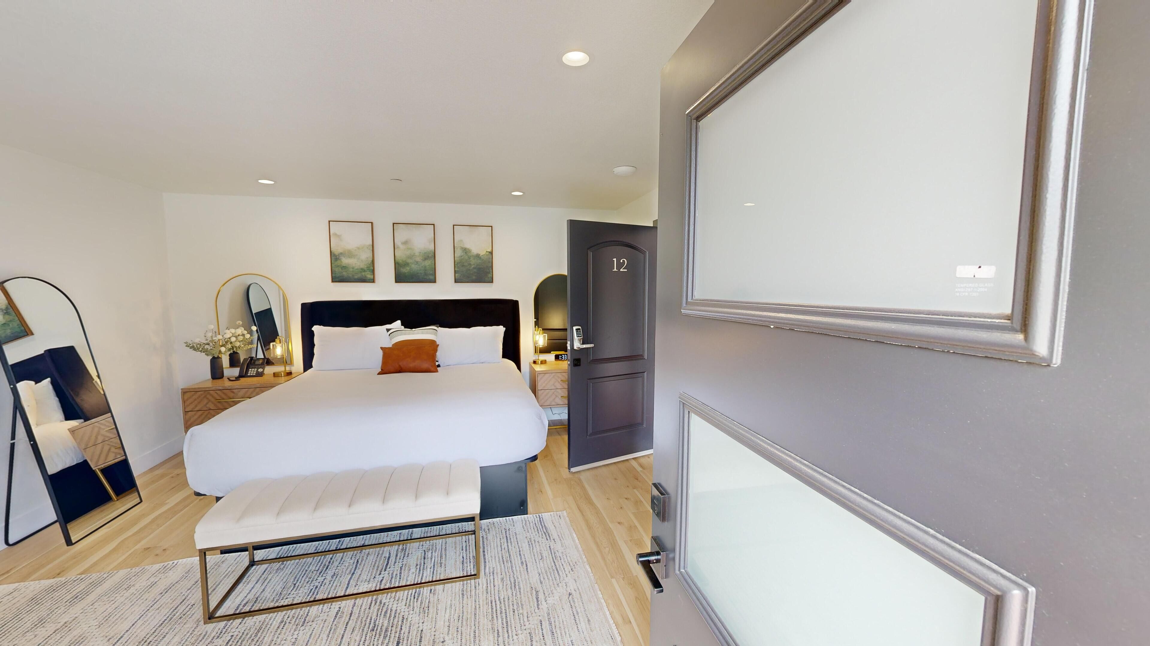 A modern hotel room with a large bed, bedside table, mirror, and artwork on the wall.