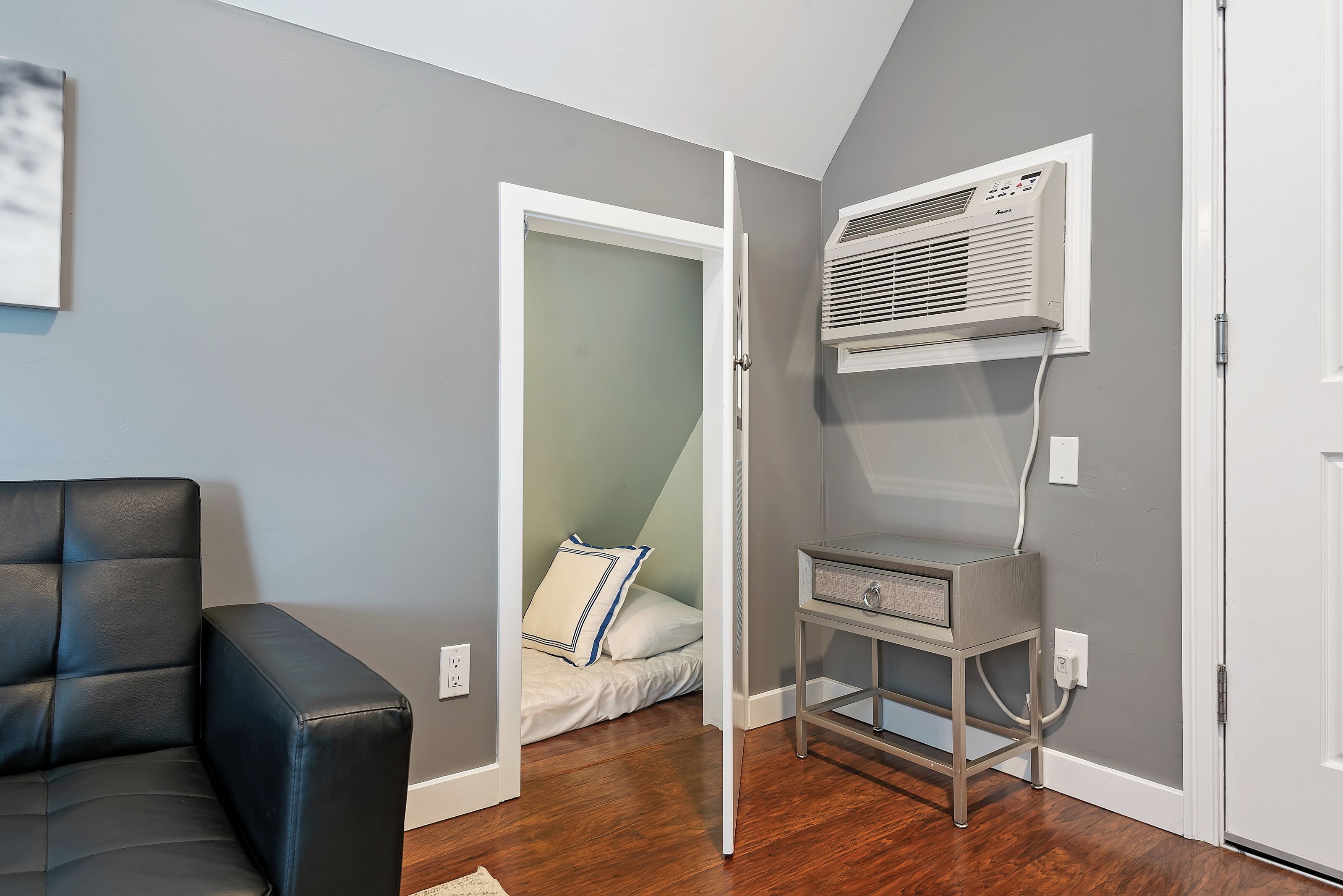 A room with a bed, a black leather sofa, a wall-mounted air conditioner, and a small table with drawers.