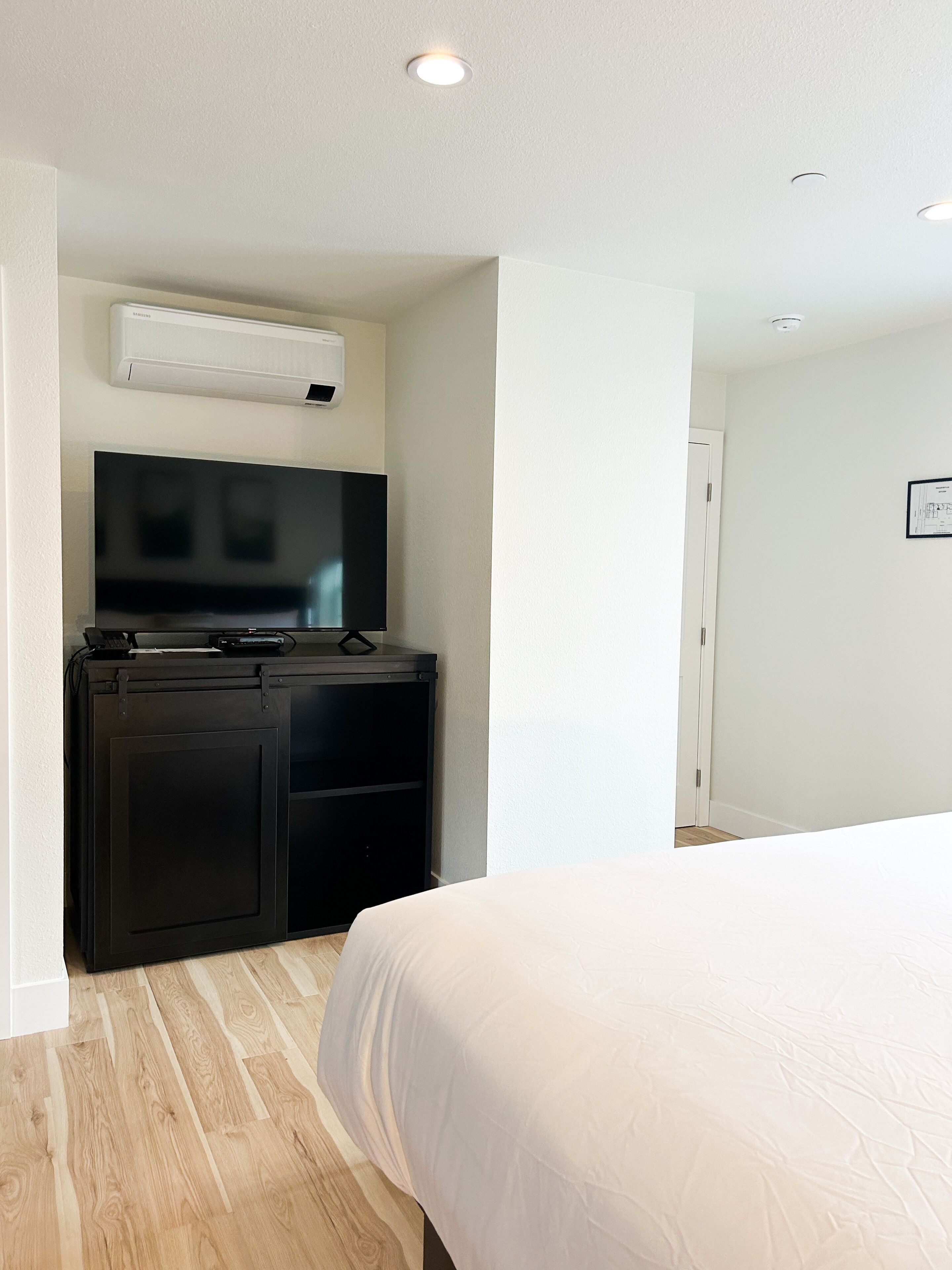 A hotel room with a bed, a desk with a flat-screen TV, and an air conditioning unit mounted on the wall.
