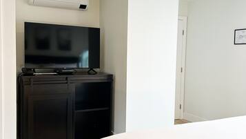 A hotel room with a bed, a desk with a flat-screen TV, and an air conditioning unit mounted on the wall.