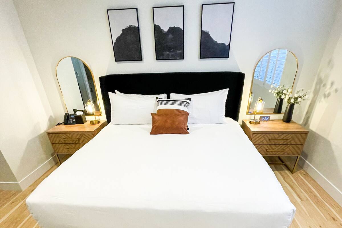 Ein modernes Hotelzimmer mit einem großen Bett, zwei Nachttischen inklusive Lampen, einem Schminkspiegel und drei gerahmten Bildern an der Wand.