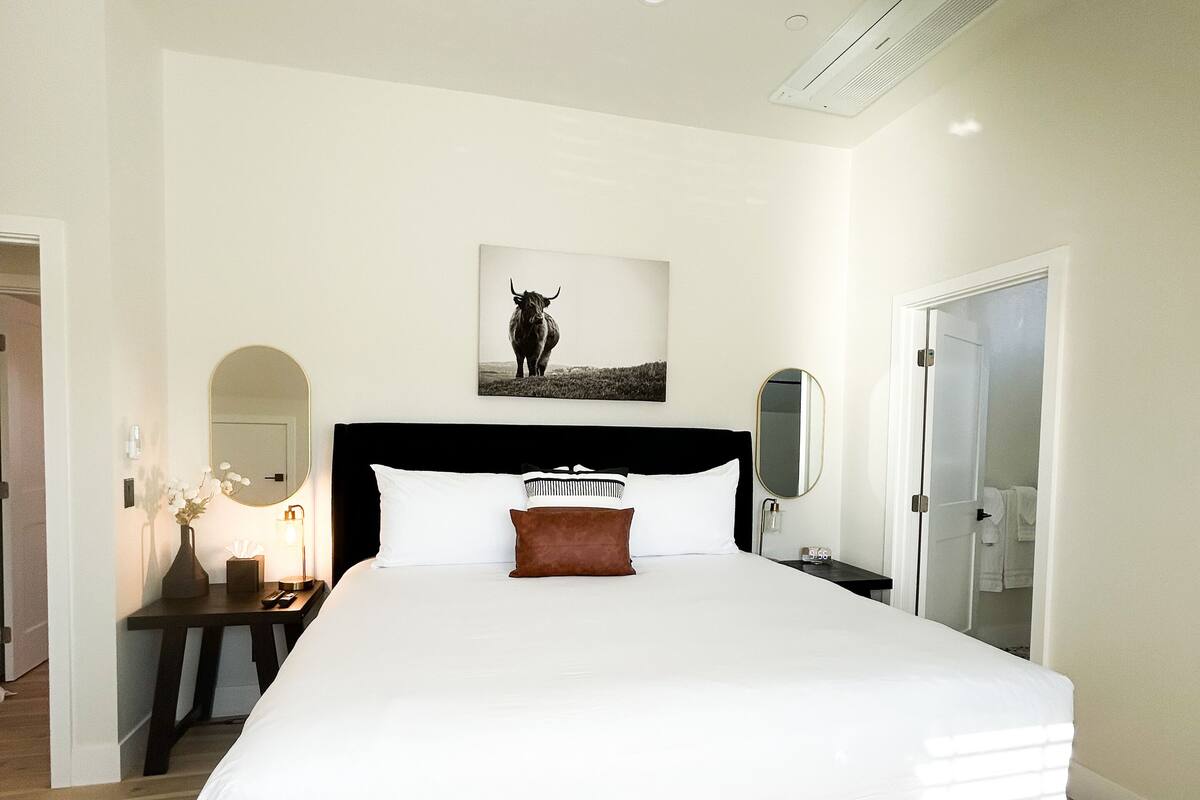 Ein modernes Schlafzimmer mit einem großen Bett, einem Nachttisch mit Lampe, einem Spiegel und einem gerahmten Bild an der Wand.