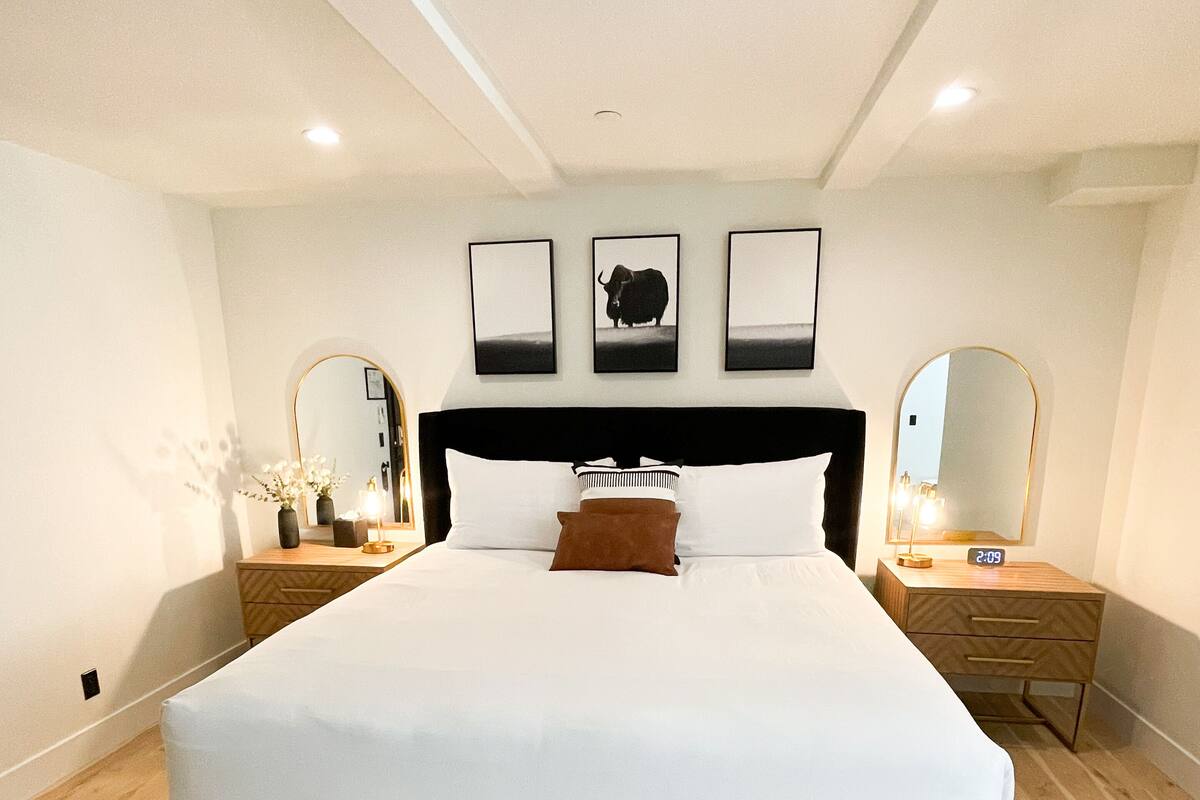 Ein modernes Schlafzimmer mit einem großen Bett, einer Bank, zwei Nachttischen, einem Beistelltisch, einem Spiegel und drei gerahmten Bildern an der Wand.