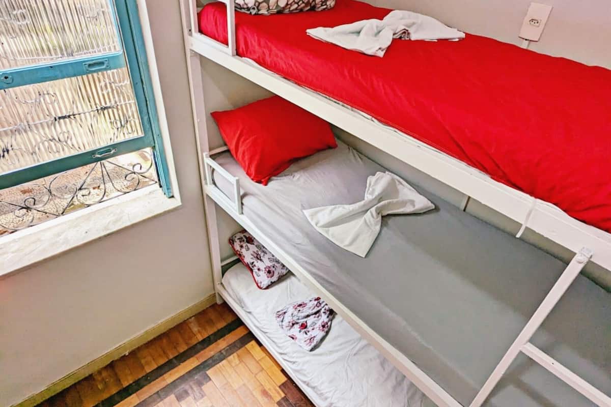 A bunk bed with red bedding and a patterned pillow, a window with a metal grill, and a herringbone-patterned wooden floor.