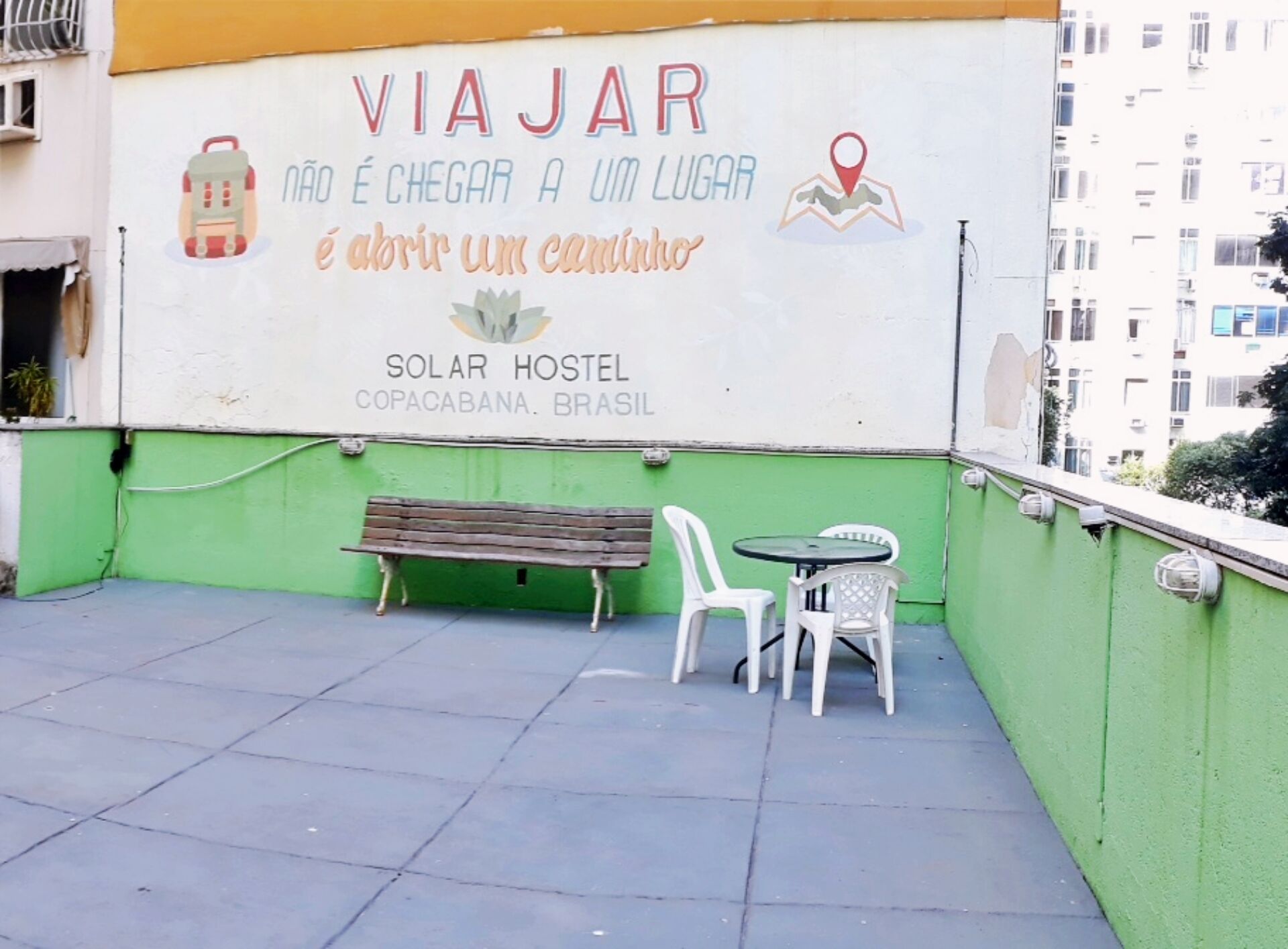 Un hostal llamado 'VIAJAR' con una pared exterior verde y blanca, un banco y dos sillas blancas.