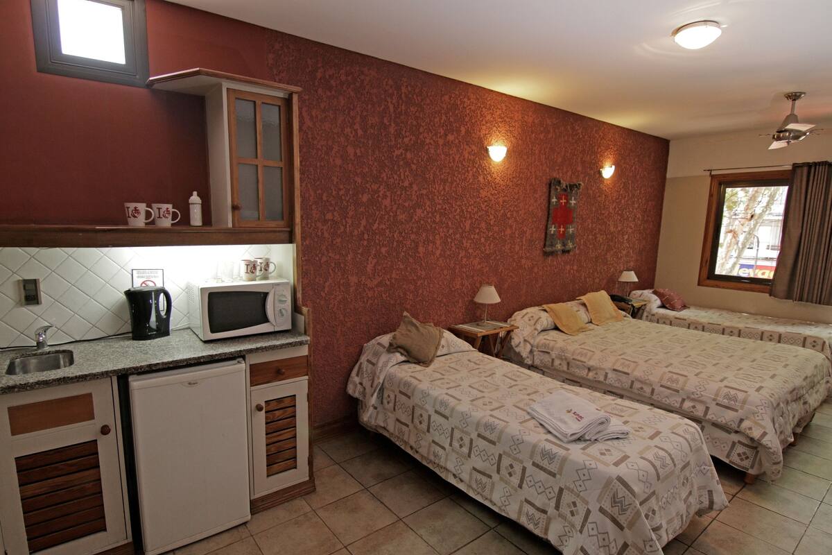 A compact hotel room with a kitchenette, two beds with patterned bedding, a small window, and a ceiling fan.