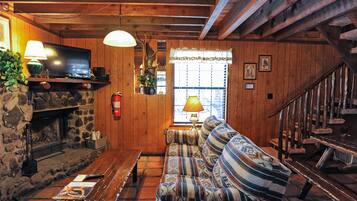 A cozy cabin interior with a stone fireplace, wooden walls, a sofa, a television, and a staircase.