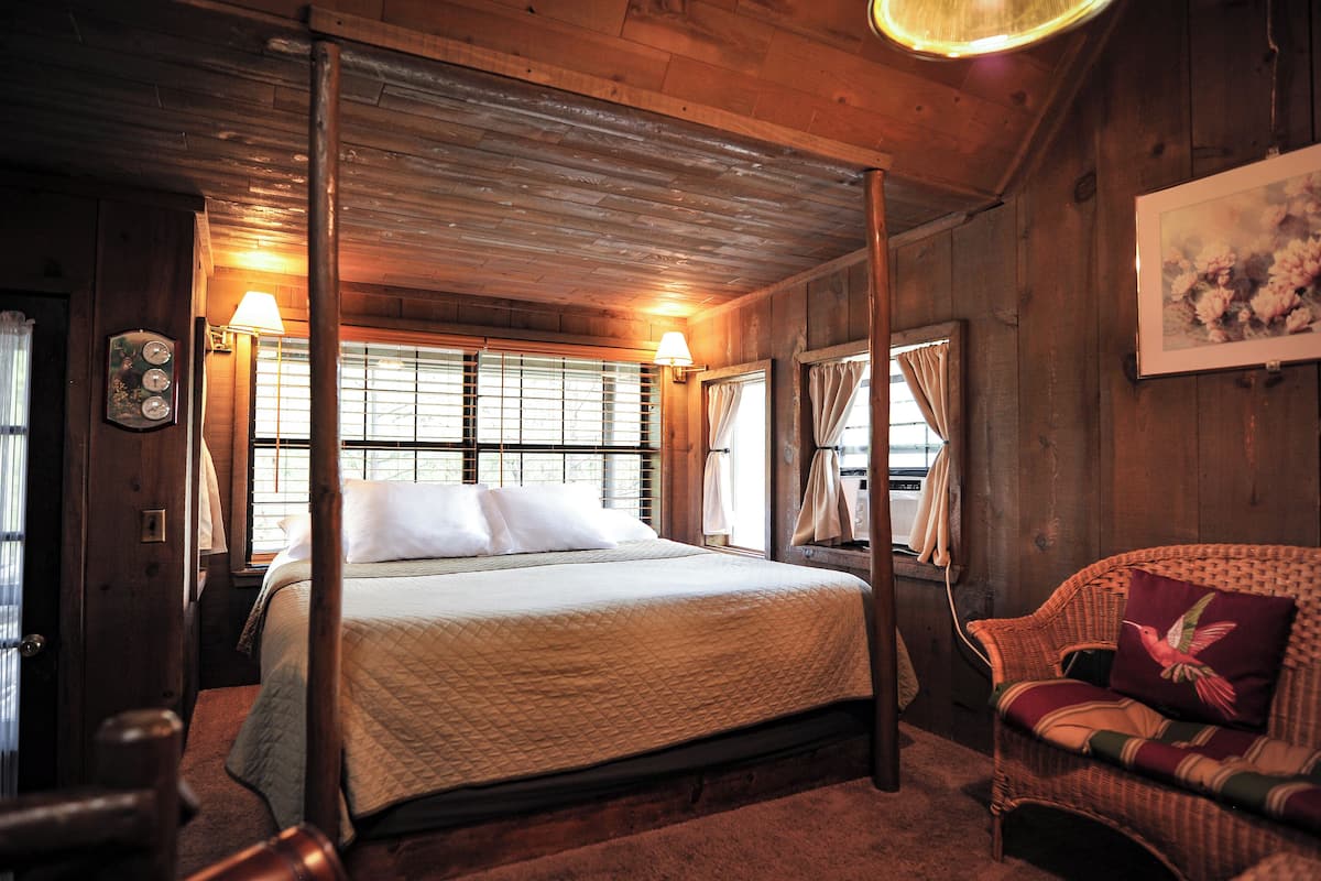 A four-poster bed with a quilted bedspread, a wicker chair with a cushion, and a framed floral painting on the wall.