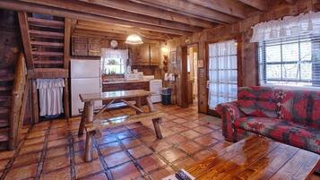 Interior rústico de una cabaña con una mesa de comedor de madera, un sofá y una zona de cocina con refrigerador y microondas.
