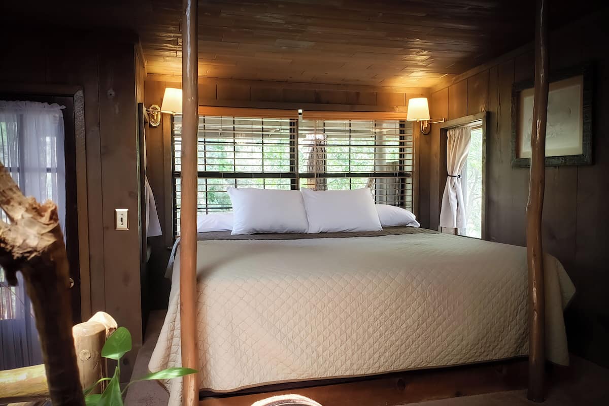 A wooden four-poster bed with a quilted mattress, two white pillows, and a window with blinds.