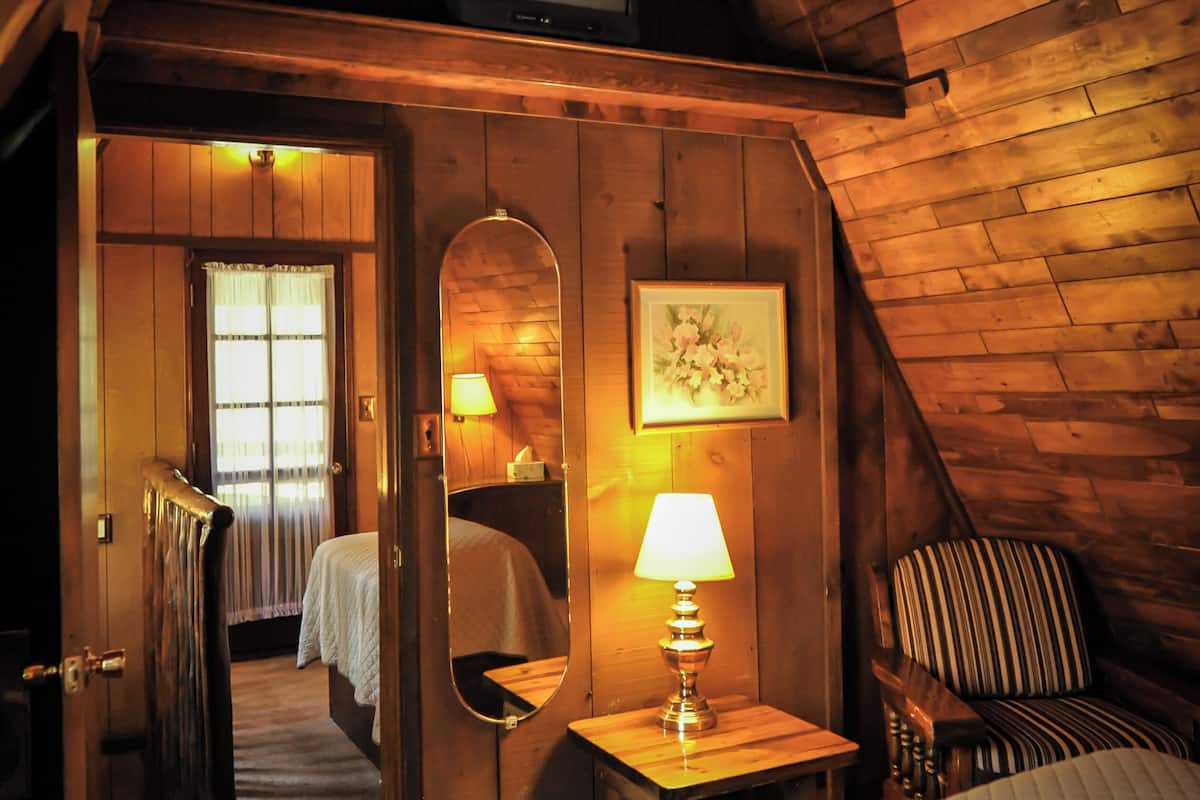 A cozy attic bedroom with a bed, a chair, a small table with a lamp, a mirror, and a television.