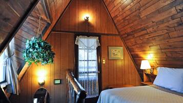 A cozy attic bedroom with wooden walls, a bed, a hanging plant, a window with lace curtains, and a nightstand with a lamp.