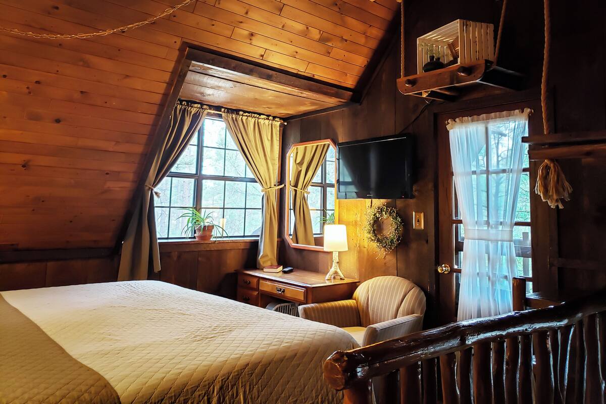 A cozy room with a bed, a desk with a lamp, a chair, a TV, and a window with curtains.