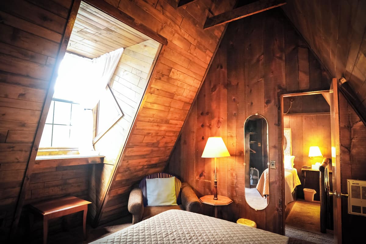 A cozy attic bedroom with a sloped ceiling, a bed, a chair, a small table, and a lamp.