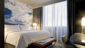 A hotel room with a large bed, a desk with a chair, a bench, and a large window with curtains.